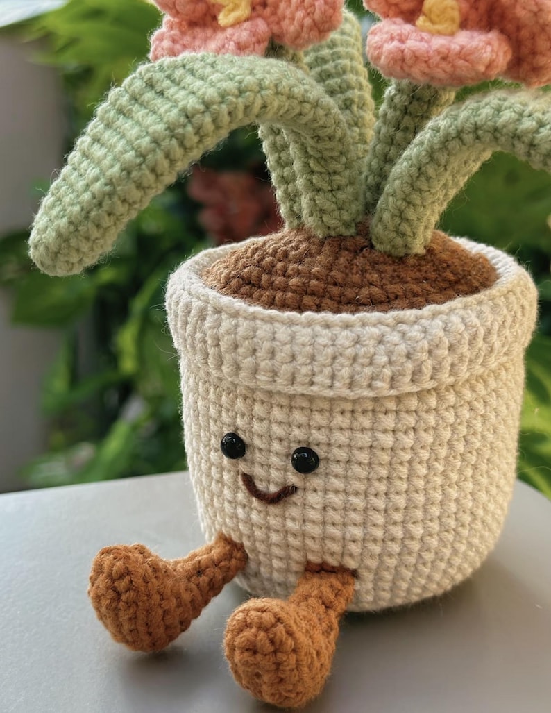 Puede incluir: Una planta de ganchillo en una maceta blanca con una cara sonriente y piernas marrones. La planta tiene tallos verdes y flores rosas. La maceta est&aacute; llena de hilo marr&oacute;n, que se asemeja a tierra. El fondo est&aacute; borroso, con follaje verde.