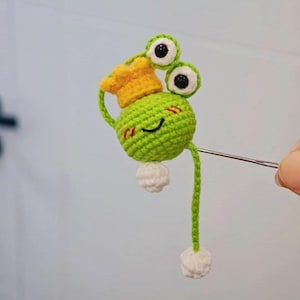 Könnte beinhalten: Eine handgefertigte gehäkelte Froschfigur, hauptsächlich grün mit weißen Akzenten und einer gelben Krone. Es hat große Wackelaugen und ein lächelndes Gesicht. Der Frosch ist an einer Metallnadel befestigt.