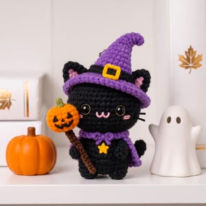 May include: A black crocheted cat plush wearing a purple witch hat with a yellow buckle and a purple cape with a yellow star. The cat holds a small orange crocheted pumpkin on a brown stick. It has black eyes, pink cheeks, and a pink stitched mouth.