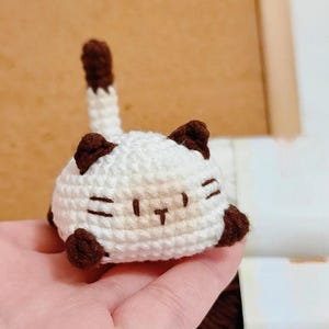 May include: A small, handmade crochet cat. The cat is white with brown detailing on the ears, paws, and tail. The face features stitched details. The figurine has a rounded shape and is small in size.