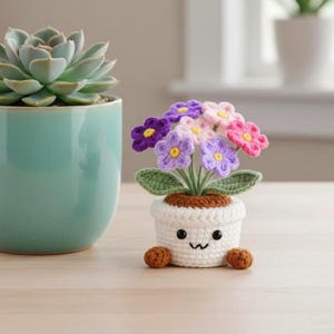 Op de afbeelding: Een gehaakte bloempot met een lachend gezicht, met een boeket van paarse, roze en gele bloemen. De pot is wit met bruine accenten en kleine bruine ballen als voetjes. Links staat een turquoise keramische pot met een vetplant.
