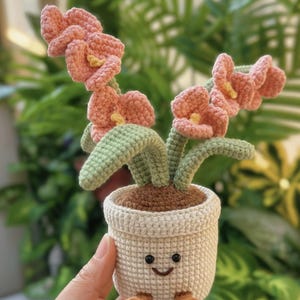 Puede incluir: Una planta de ganchillo con flores rosas, hojas verdes y una maceta blanca sonriente con pies marrones. La planta hecha a mano es un art&iacute;culo decorativo &uacute;nico.