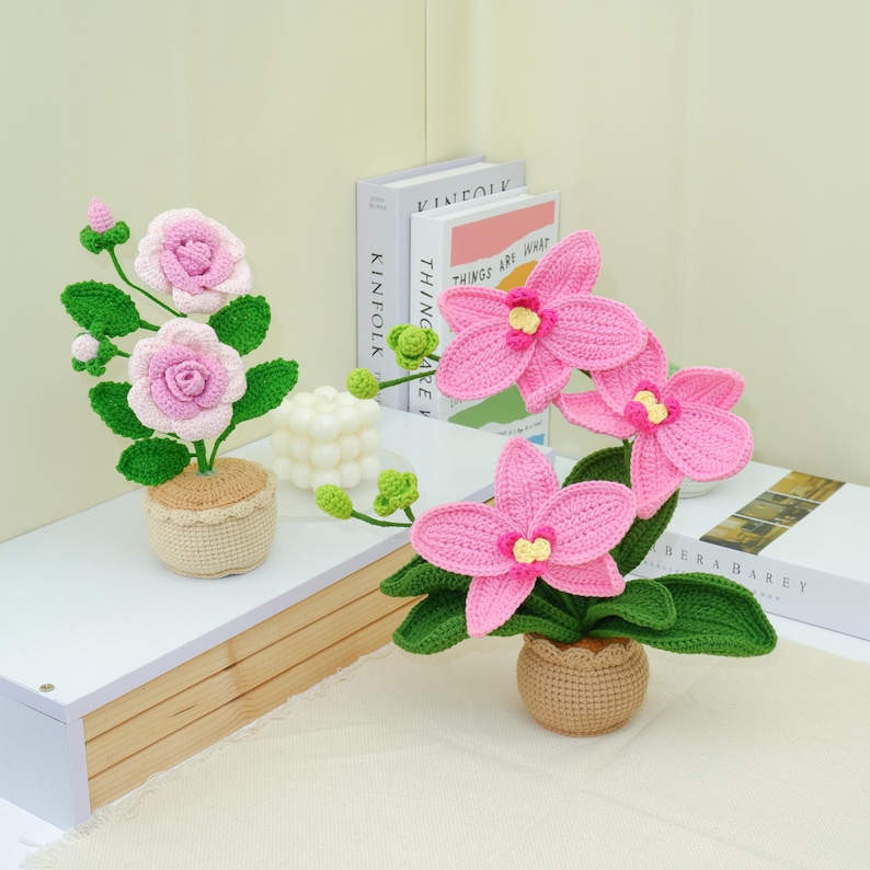 May include: Crocheted pink orchid and rose flowers in small pots. The flowers have green leaves and stems. The pots are a light tan color. The flowers are on a white shelf with books in the background.