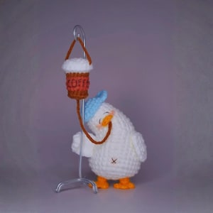以下が含まれることがあります： 青い帽子をかぶり、オレンジ色の足と胸に小さな黒い十字架が縫い付けられた、手作りのかぎ針編みのカモメの置物。カモメは「COFFEE」の文字が入ったコーヒーカップを持っています。コーヒーカップは金属製のスタンドから吊り下げられています。
