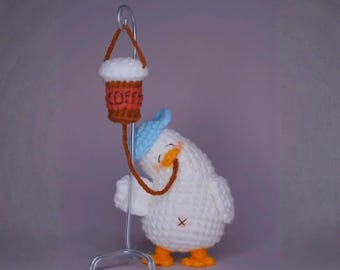 May include: A handmade crochet seagull figurine wearing a blue hat, with orange feet and a small black cross stitched on its chest. The seagull is holding a coffee cup with the word "COFFEE" on it. The coffee cup is suspended from a metal stand.