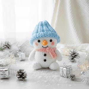 May include: A handmade crochet snowman wearing a light blue hat and a pink scarf. The snowman features a carrot nose, rosy cheeks, and black button eyes. The scene includes silver gift boxes, pine cones, and star-shaped lights, evoking a winter theme.