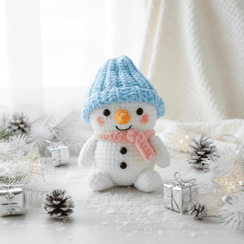 May include: A handmade crochet snowman wearing a light blue hat and a pink scarf. The snowman features a carrot nose, rosy cheeks, and black button eyes. The scene includes silver gift boxes, pine cones, and star-shaped lights, evoking a winter theme.