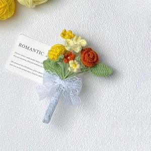 Puede incluir: Un ramo de flores de ganchillo hecho a mano con flores amarillas, naranjas y rojas, hojas verdes y un lazo de encaje blanco. La tarjeta dice "ROMANTIC".