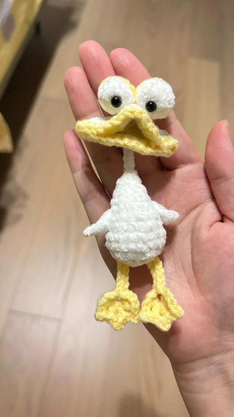 May include: A crocheted duck toy, featuring a white body, yellow beak, and feet. The duck has large white eyes with black pupils. The duck's body is fluffy white, and the beak and feet are bright yellow. A charming, handcrafted item.