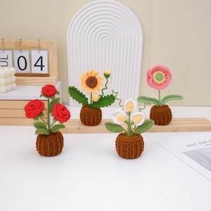 Puede incluir: Cuatro arreglos florales de ganchillo hechos a mano en macetas marrones pequeñas. Las flores incluyen rosas rojas, un girasol amarillo, calas blancas y amarillas y una flor rosa. Las flores están sobre una repisa de madera.