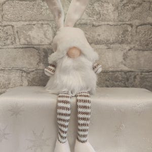 May include: A white Easter gnome with long bunny ears, a fluffy white beard, and a pink nose. The gnome's legs are striped brown and white, and it is sitting on a white surface. The background is a light grey brick wall.