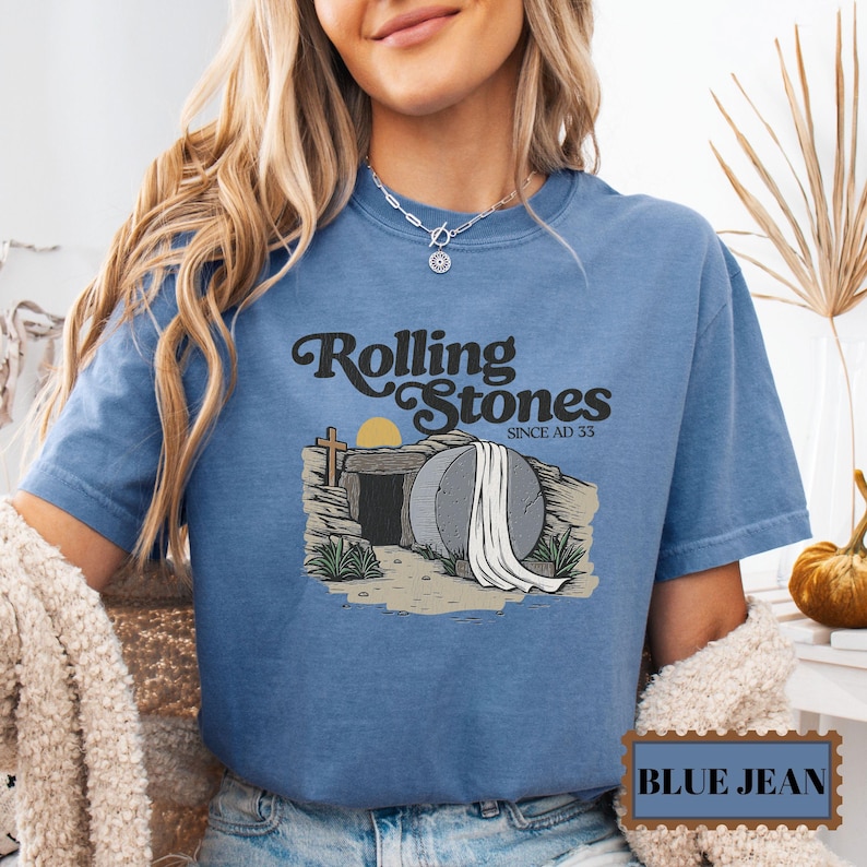 May include: A blue jean t-shirt with a graphic of a stone tomb with a white sheet on top. The text "Rolling Stones Since AD 33" is above the tomb.