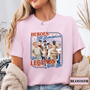 May include: A light pink t-shirt featuring a graphic with the text "HEROES Get Remembered But LEGENDS Never Die." The graphic includes an image of a group of boys. The word "BLOSSOM" is in a rectangular box at the bottom right.