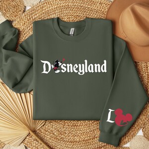 Puede incluir: Una sudadera verde oliva con la palabra "Disneyland" en escritura blanca. Un gr&aacute;fico de Mickey Mouse est&aacute; en la parte delantera, y la manga presenta la palabra "I" y una cabeza de Mickey Mouse. Un sombrero marr&oacute;n descansa en el fondo.