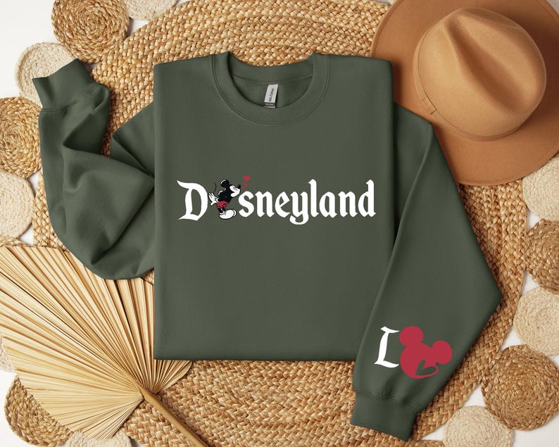 Puede incluir: Una sudadera verde oliva con la palabra "Disneyland" en escritura blanca. Un gr&aacute;fico de Mickey Mouse est&aacute; en la parte delantera, y la manga presenta la palabra "I" y una cabeza de Mickey Mouse. Un sombrero marr&oacute;n descansa en el fondo.