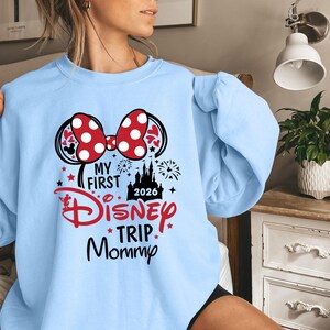 May include: Light blue sweatshirt with a graphic featuring Minnie Mouse ears, a red polka dot bow, and the text "My First Disney Trip Mommy 2026". The design includes a castle, stars, and fireworks.