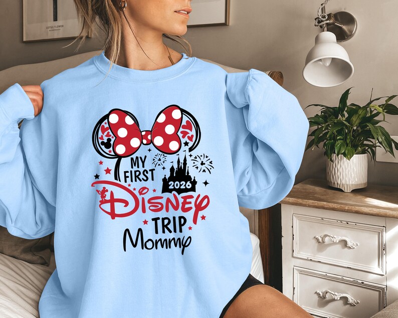 May include: Light blue sweatshirt with a graphic featuring Minnie Mouse ears, a red polka dot bow, and the text "My First Disney Trip Mommy 2026". The design includes a castle, stars, and fireworks.