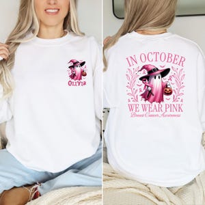 May include: White sweatshirt with a pink ghost wearing a witch hat and holding a pumpkin. The text "In October We Wear Pink Breast Cancer Awareness" is on the back of the sweatshirt. The front of the sweatshirt has a smaller pink ghost with the name "Olivia" underneath.