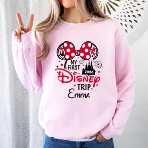 May include: A light pink sweatshirt with a graphic featuring a Minnie Mouse bow with red polka dots, the text "My First Disney Trip 2026 Emma", and fireworks and castle illustrations.
