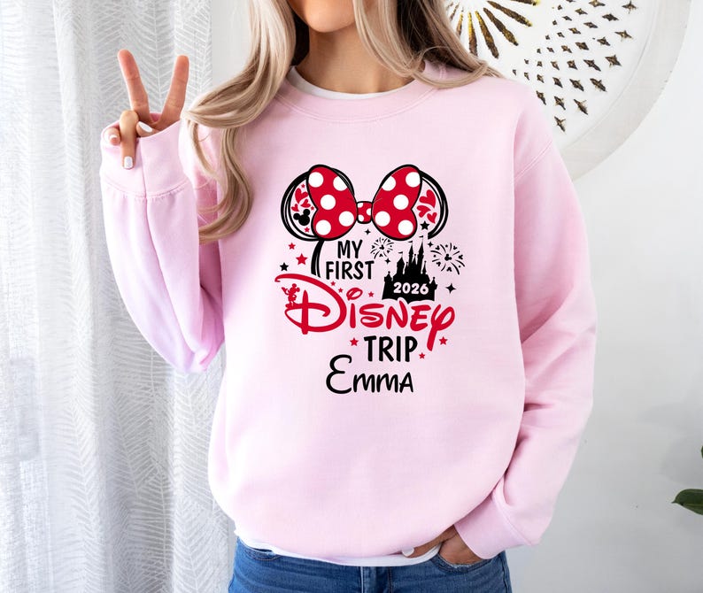 May include: A light pink sweatshirt with a graphic featuring a Minnie Mouse bow with red polka dots, the text "My First Disney Trip 2026 Emma", and fireworks and castle illustrations.