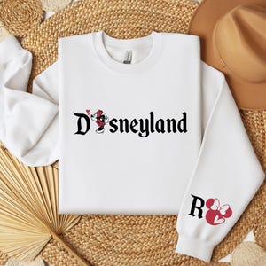 Puede incluir: Sudadera blanca con la palabra "Disneyland" en escritura negra, con gr&aacute;ficos de Mickey Mouse y Minnie Mouse. La manga derecha tiene la letra "R" y un gr&aacute;fico de Minnie Mouse. La sudadera est&aacute; sobre un fondo tejido.