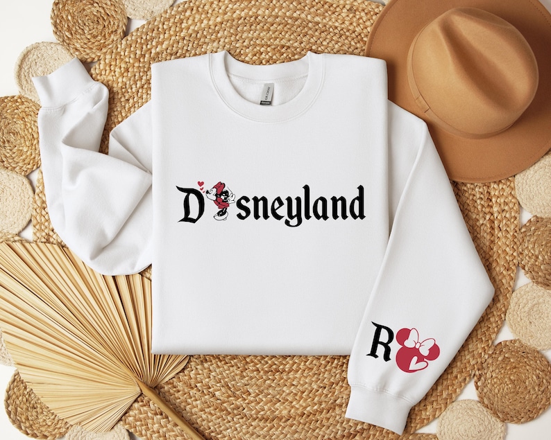 Puede incluir: Sudadera blanca con la palabra "Disneyland" en escritura negra, con gr&aacute;ficos de Mickey Mouse y Minnie Mouse. La manga derecha tiene la letra "R" y un gr&aacute;fico de Minnie Mouse. La sudadera est&aacute; sobre un fondo tejido.