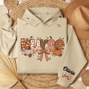 May include: A beige hoodie with a fall-themed design that says "Nurse" with pumpkins and leaves. The hoodie has a personalized name on the sleeve that says "Olivia".