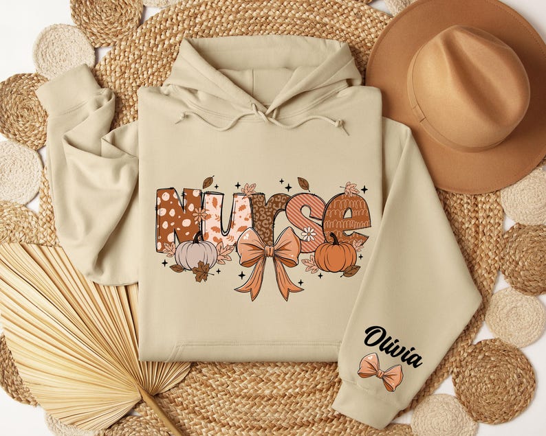May include: A beige hoodie with a fall-themed design that says "Nurse" with pumpkins and leaves. The hoodie has a personalized name on the sleeve that says "Olivia".