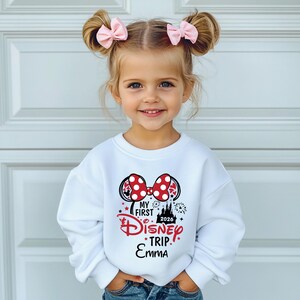 May include: A young girl wearing a white sweatshirt with a Minnie Mouse-themed design. The design includes a red polka-dot bow, the words "My First Disney Trip" and the year 2026. The girl is also wearing pink bows in her hair.
