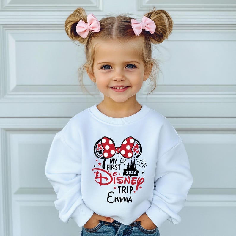 May include: A young girl wearing a white sweatshirt with a Minnie Mouse-themed design. The design includes a red polka-dot bow, the words "My First Disney Trip" and the year 2026. The girl is also wearing pink bows in her hair.