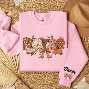 May include: A pink sweatshirt with a graphic of the word "Nurse" in fall-themed patterned letters, a bow, pumpkins, and leaves. The word "Olivia" is written on the sleeve with a small bow.