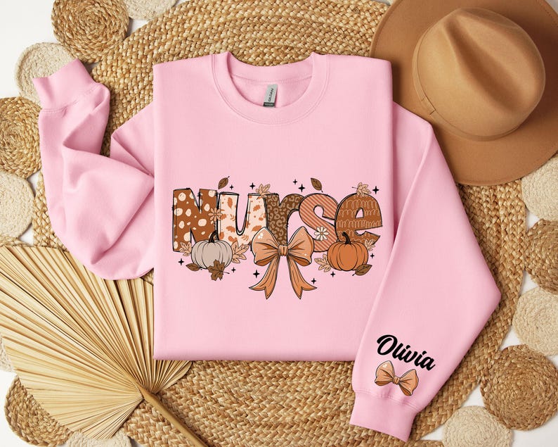May include: A pink sweatshirt with a graphic of the word "Nurse" in fall-themed patterned letters, a bow, pumpkins, and leaves. The word "Olivia" is written on the sleeve with a small bow.