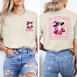 May include: A light beige t-shirt with a pink ghost wearing a witch hat and holding a pumpkin. The text "In October We Wear Pink" and "Breast Cancer Awareness" is on the back of the shirt. The front of the shirt has the text "Olivia" and a smaller pink ghost.