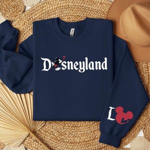 Puede incluir: Sudadera azul marino con la palabra "Disneyland" en escritura blanca, con un dibujo de Mickey Mouse. La manga izquierda muestra la palabra "Love" con un dibujo de la cabeza de Mickey Mouse en rojo. Un sombrero marr&oacute;n claro descansa en la parte superior derecha.