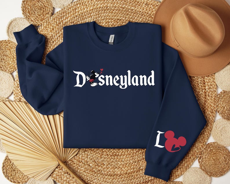 Puede incluir: Sudadera azul marino con la palabra "Disneyland" en escritura blanca, con un dibujo de Mickey Mouse. La manga izquierda muestra la palabra "Love" con un dibujo de la cabeza de Mickey Mouse en rojo. Un sombrero marr&oacute;n claro descansa en la parte superior derecha.