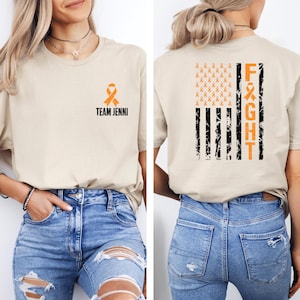 May include: Beige t-shirt with an orange ribbon and the text "TEAM JENNI" on the front. The back features a distressed American flag design with orange ribbons and the word "FIGHT" in orange.