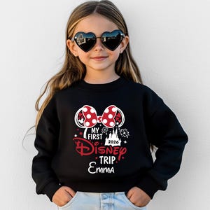 May include: A young girl wearing heart-shaped sunglasses and a black sweatshirt. The sweatshirt features a Minnie Mouse bow design with red polka dots and the text "My First Disney Trip 2026 Emma" in red and white.
