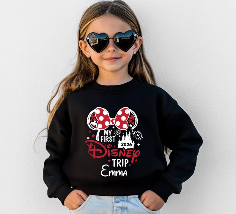 May include: A young girl wearing heart-shaped sunglasses and a black sweatshirt. The sweatshirt features a Minnie Mouse bow design with red polka dots and the text "My First Disney Trip 2026 Emma" in red and white.