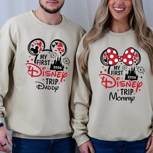 May include: Beige sweatshirts with Disney-themed graphics. One features Mickey Mouse ears and the text "My First Disney Trip Daddy 2026." The other has a Minnie Mouse bow and reads "My First Disney Trip Mommy 2026."