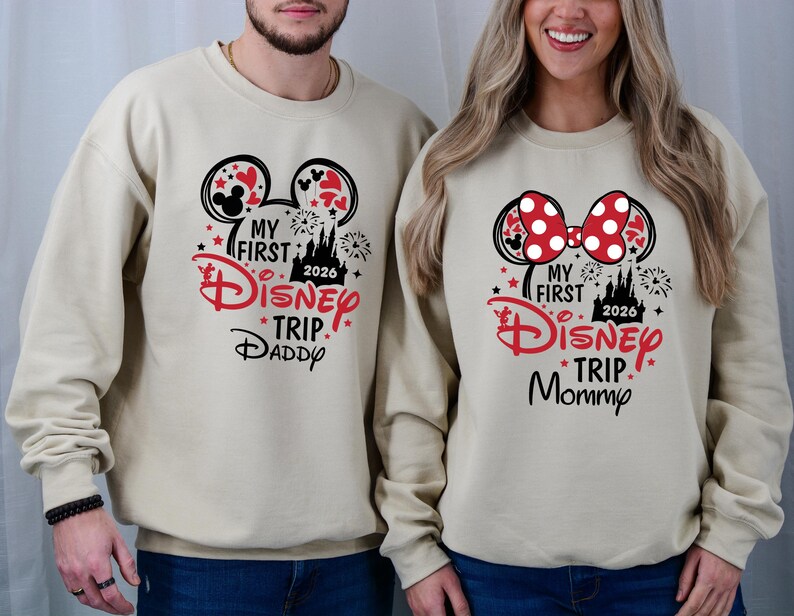 May include: Beige sweatshirts with Disney-themed graphics. One features Mickey Mouse ears and the text "My First Disney Trip Daddy 2026." The other has a Minnie Mouse bow and reads "My First Disney Trip Mommy 2026."