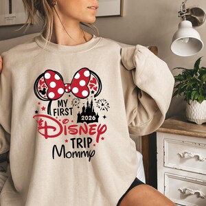 May include: Beige sweatshirt with a graphic featuring a red and white polka dot bow, Mickey Mouse ears, and the text "My First Disney Trip Mommy 2026". The design includes a castle and fireworks.
