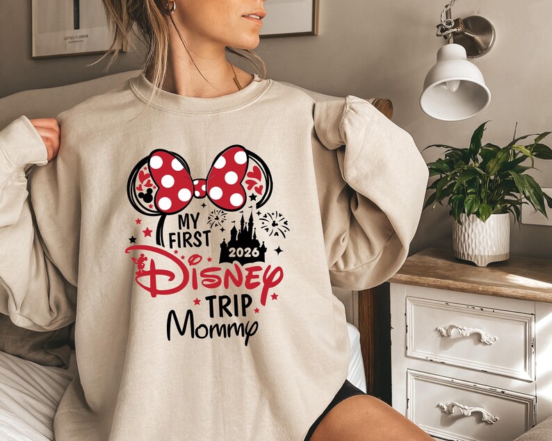 May include: Beige sweatshirt with a graphic featuring a red and white polka dot bow, Mickey Mouse ears, and the text "My First Disney Trip Mommy 2026". The design includes a castle and fireworks.