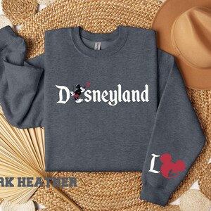 Puede incluir: Sudadera gris oscuro con la palabra "Disneyland" en escritura blanca, con Mickey Mouse. La manga izquierda tiene una cabeza de Mickey Mouse roja y la letra "L".