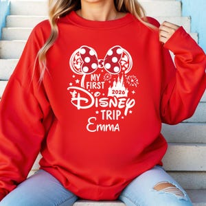 May include: A red sweatshirt with a white graphic featuring a Minnie Mouse bow, the words "My First Disney Trip 2026 Emma", and Disney-themed illustrations. The sweatshirt is a solid red color.