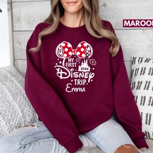 May include: Maroon sweatshirt with a white graphic featuring Minnie Mouse ears with a red polka dot bow, the text "MY FIRST Disney TRIP" and the name "Emma". The year "2026" is also included.