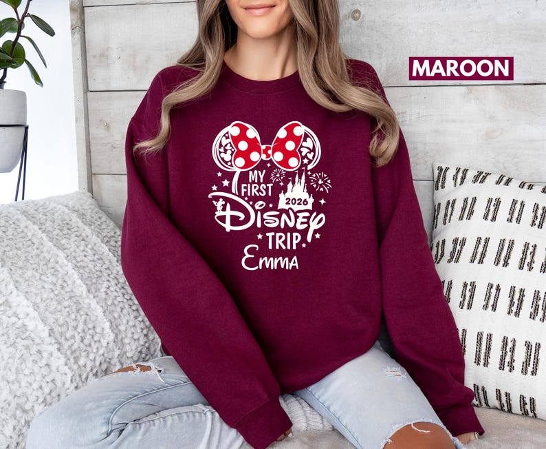 May include: Maroon sweatshirt with a white graphic featuring Minnie Mouse ears with a red polka dot bow, the text "MY FIRST Disney TRIP" and the name "Emma". The year "2026" is also included.