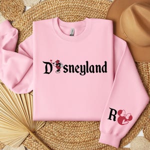 Puede incluir: Una sudadera rosa claro con la palabra "Disneyland" en fuente g&oacute;tica negra, con un gr&aacute;fico de Minnie Mouse. La manga derecha muestra la letra "R" y una cabeza de Minnie Mouse con un lazo. La sudadera est&aacute; hecha de un material suave.