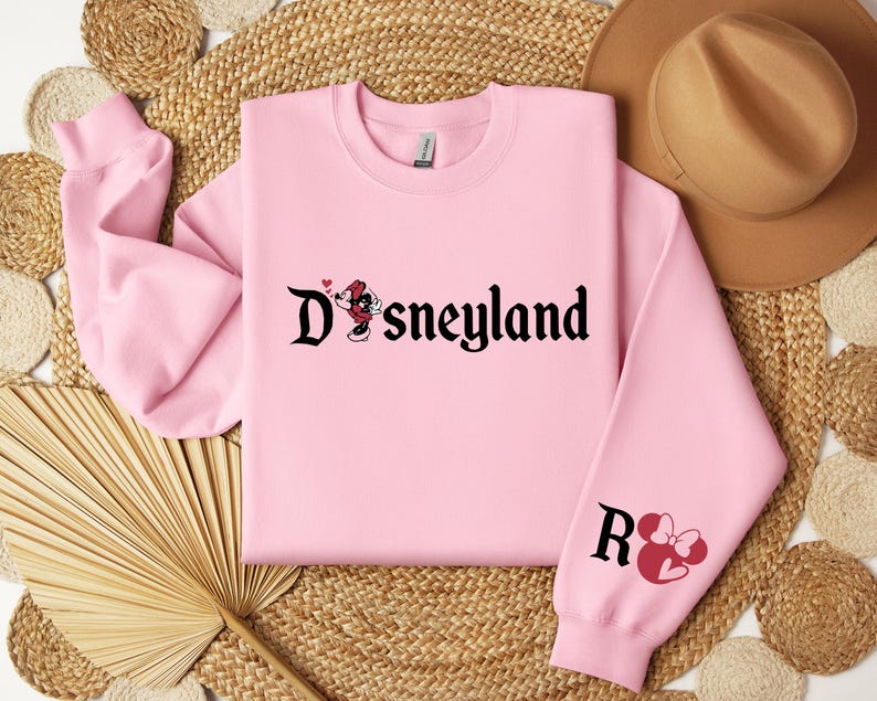 Puede incluir: Una sudadera rosa claro con la palabra "Disneyland" en fuente g&oacute;tica negra, con un gr&aacute;fico de Minnie Mouse. La manga derecha muestra la letra "R" y una cabeza de Minnie Mouse con un lazo. La sudadera est&aacute; hecha de un material suave.