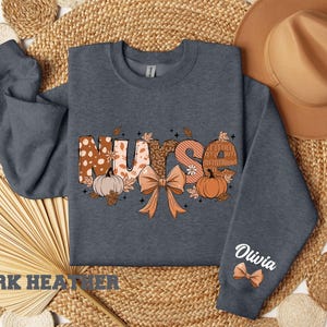 May include: Dark heather grey sweatshirt with a graphic of the word "Nurse" in fall colors with pumpkins, leaves, and a bow. The text "Olivia" is on the sleeve.