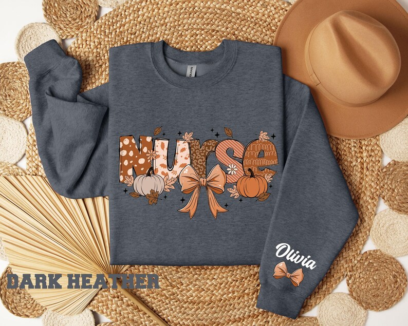 May include: Dark heather grey sweatshirt with a graphic of the word "Nurse" in fall colors with pumpkins, leaves, and a bow. The text "Olivia" is on the sleeve.